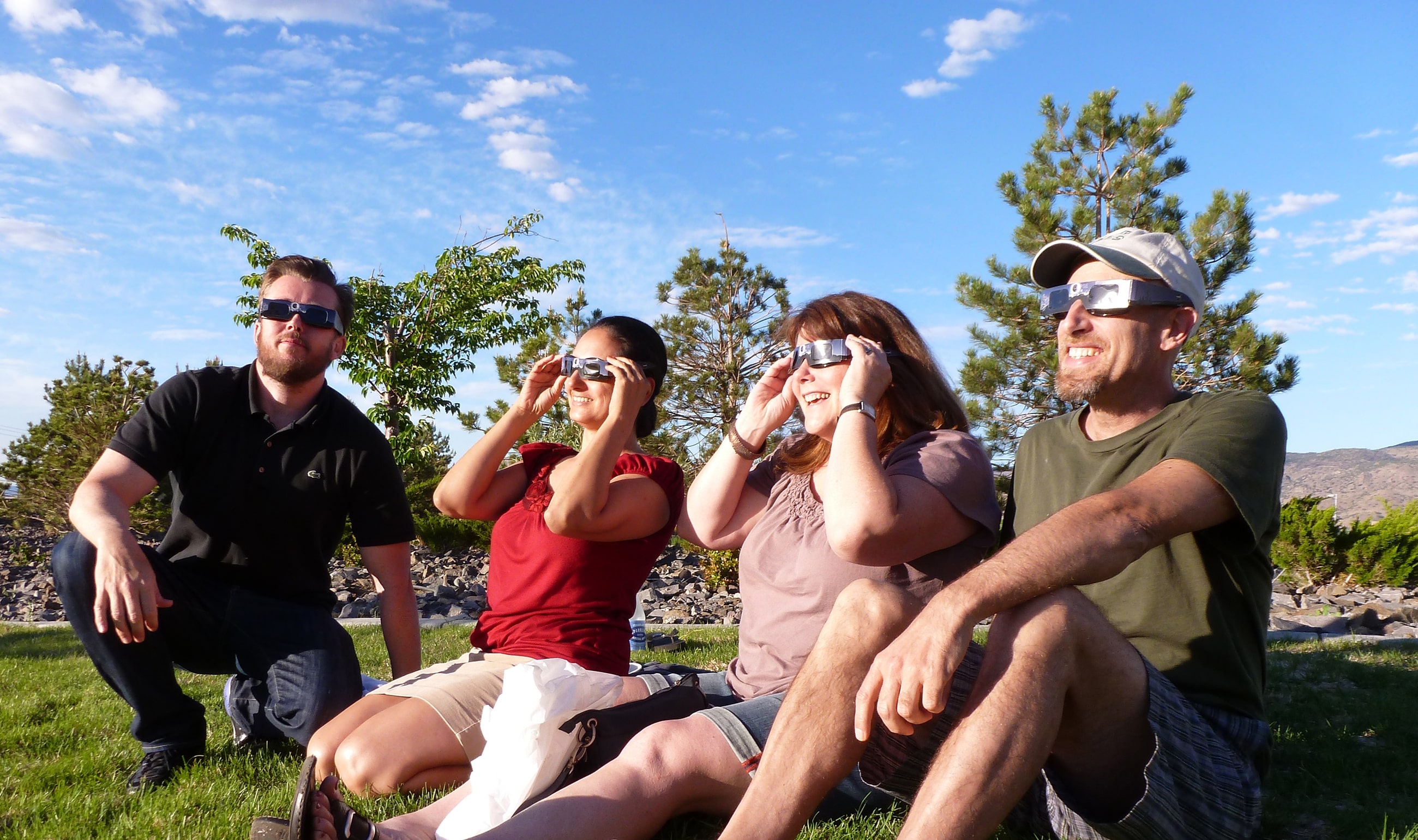 A group of 4 eclipse watchers wearing eclipse-viewing glasses (glasses that filter the Sun’s harmful UV rays, allowing you to look right at it) look up at the sun in the sky. They are smiling and sitting on a green lawn with trees behind them.