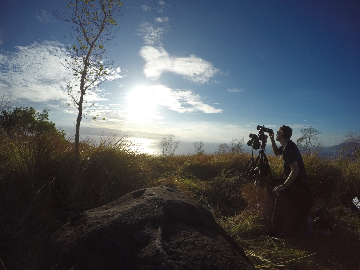 A man surrounded by tall, mostly dead, grass peers through his camera, which sits on a tripod and is pointed at the sky where the sun is shining. 