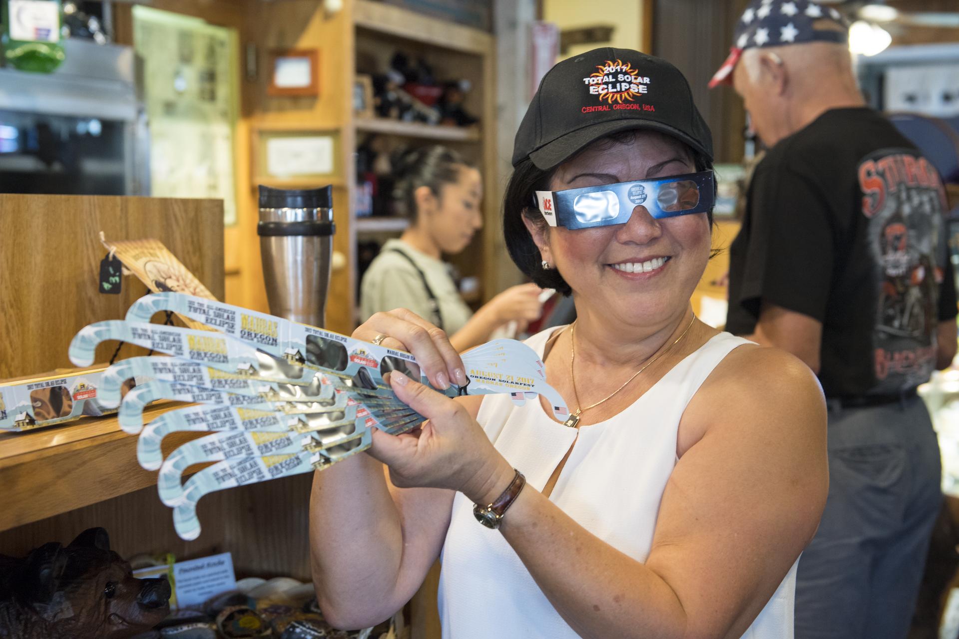 A smiling woman in a store wearing a hat that says “2017 Total Solar Eclipse” holds up a handful of eclipse-viewing glasses fanned out so they can all be seen. In the background, a man stands at a counter and pays for his items. 